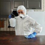 A person in protective clothing disinfects a kitchen counter indoors, focusing on safety and cleanliness. disinfeksi